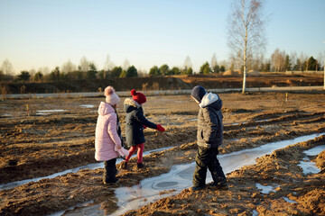 Group of children playing with thin ice puddles formed on the frozen soil in winter. Kids having fun in winter.
