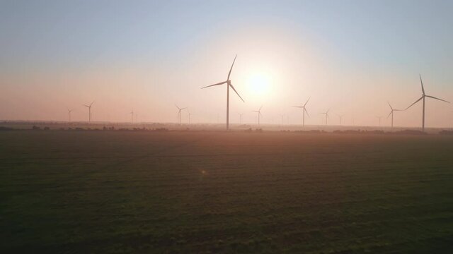 Windmills with rotating wings at sunset or sunrise. Technology for wind energy, renewable energy source, earth care. Wind power station, drone video, moving toward the horizon, 4k