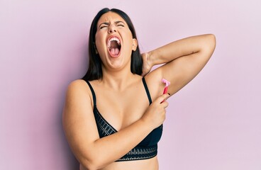 Beautiful brunette woman shaving armpit hair with razor angry and mad screaming frustrated and furious, shouting with anger looking up. - obrazy, fototapety, plakaty