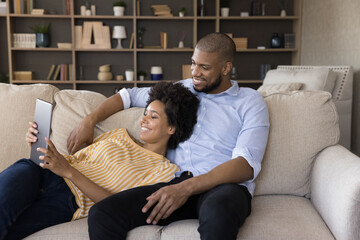 Happy African American married couple watching media content on tablet computer together, making video call, using digital device, resting on sofa in living room, talking, smiling, laughing