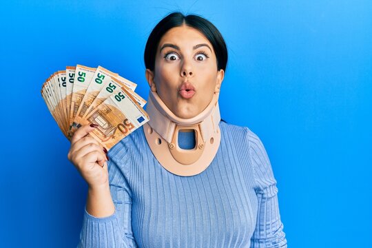 Beautiful brunette woman wearing cervical collar holding insurance money scared and amazed with open mouth for surprise, disbelief face