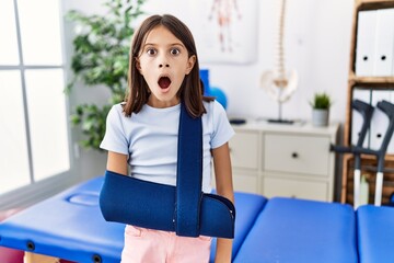 Young hispanic girl wearing arm on sling at rehabilitation clinic scared and amazed with open mouth for surprise, disbelief face