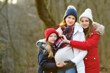 Fototapeta premium Young pregnant woman hugging her older daughters. Older siblings having fun with her pregnant mom outdoors. Mother and her kids spending time together.