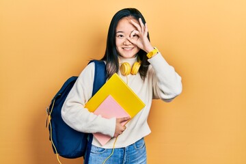 Young chinese girl holding student backpack and books doing ok gesture with hand smiling, eye looking through fingers with happy face.