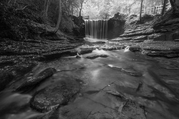 Black and white Epic beautiful Autumn landscape image of Nant Mill waterfall in Wales with glowing sunlight through the woodland