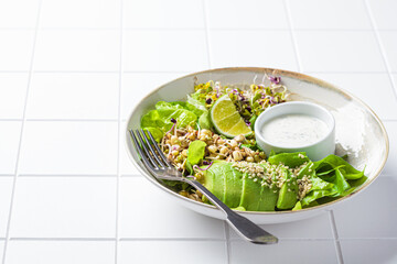 Green salad with avocado, sprouts, hemp seeds and yogurt dressing, white tile background.