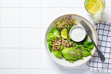 Green salad with avocado, sprouts, hemp seeds and yogurt dressing, white tile background.