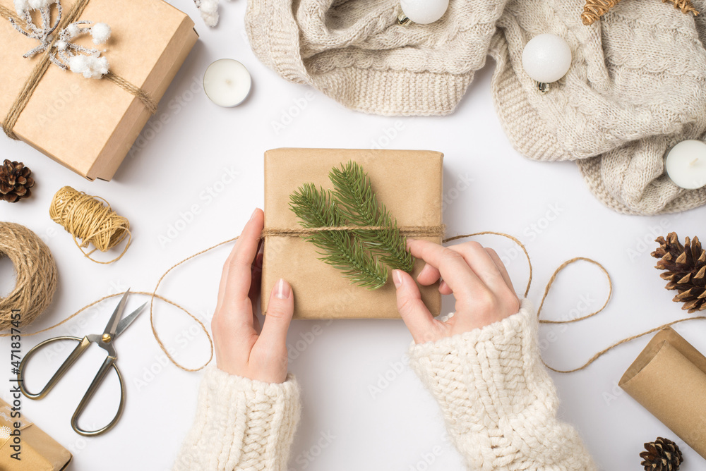 Wall mural First person top view photo of female hands in sweater decorating craft paper gift box with fir twigs knitted sweater christmas tree balls and tools for handicraft on isolated white background