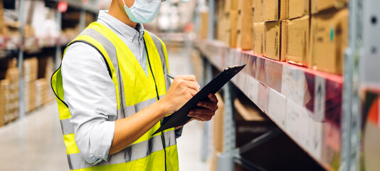 Portrait of asian engineer man in helmet in quarantine for coronavirus wearing protective mask order details checking goods and supplies on shelves with goods background in warehouse.logistic