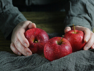 Three red apples lie on a wooden table. Hands hold them. There is a brown towel next to it.