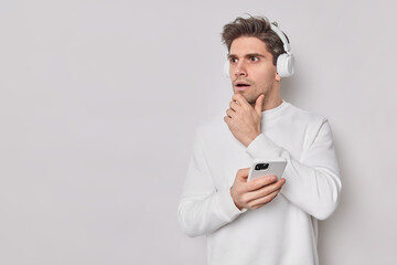 Serious shocked man holds cin and looks attentively away dressed in casual jumper listens audio track via headphones holds mobile phone isolated over white background with blank empty space.