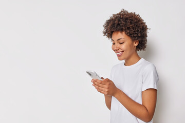 Sideways shot of happy good looking woman with curly hair uses mobile phone chats online uses application dressed in casual t shirt isolated over white background copy space for your promotion - 471841623