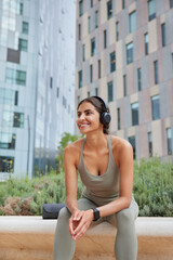 Vertical shot of sporty slim woman rests in urban setting after workout dressed in sportswear listens music via wireless headphones concentrated into distance leads actove lifestyle. Fitness training