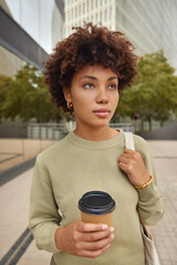 Outdoor shot of curly haired young woman in casual jumper drinks takeaway coffee carries bag...