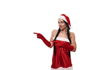 Beautiful woman in a Santa Claus carnival costume smiling cute, looking mysteriously at camera and pointing with finger on a copy space, isolated on white background. Christmas advertisement concept