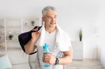 Fototapeta premium Sporty mature man holding bottle of water and sports bag, posing after domestic training and smiling, free space
