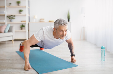 Fototapeta premium Strong senior man making strength workout, doing push ups on sports mat, exercising at home, copy space
