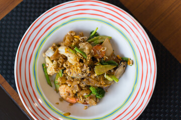 stir fried wide noodles with seafood or fried rice noodles with seafood soy sauce in white colorful rim plate on top view wooden table and placemat which consists of mussel shrimp squid and vegetable