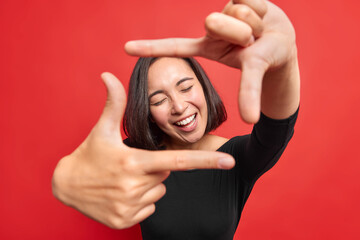 Fototapeta na wymiar Beautiful cheerful woman makes frame gesture smiles positively keeps eyes closed composes picture idea dressed in black jumper poses against vivid red background gazes at camera through hands