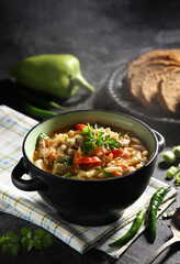 Soup, lunch. Lagman soup with noodles, vegetables, meat and fresh herbs in black bowl on the black table. Raw green pepper, cilantro, bread on black plate. Background image, copy space