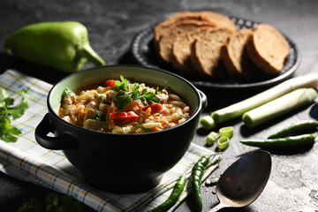 Soup, lunch. Lagman soup with noodles, vegetables, meat and fresh herbs in black bowl on the black table. Raw green pepper, cilantro, bread on black plate. Background image, copy space
