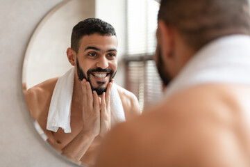 Confident young Arab guy looking in the mirror, touching beard