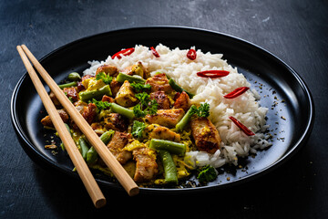 Fried chicken nuggets in curry sauce with white rice, green beans, parsley and chili peppers on black wooden table
