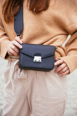 Closeup of black small handbag in woman's hand. Fall spring fashion outfit camel sweater and trendy white jeans. Fashion detail, stylish small bag.  