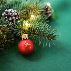 Obraz na płótnie Canvas Christmas red ball, Christmas tree, cones and garland with lights on green background. Concept: Christmas and New Year, winter holiday theme. Selective focus. Square.