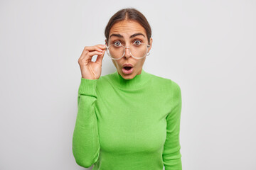 Brunette young woman wears spectacles for good vision correction and green jumper reacts on awesome news isolated over white background stares in complete disbelief. Human reactions concept.