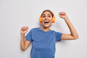 Energetic positive brunette young woman shakes arms dances with rhythm of music via headphones has upbeat mood dressed in casual blue t shirt isolated over white background. Noise reduction.