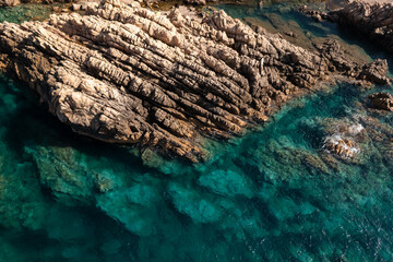 Aerial top down shot of a stone shoreline and turquoise sea water illuminated by the sun. © 24K-Production