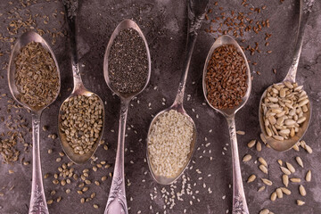 Assorted healthy seeds - sunflower, flax, chia, sesame, hemp, dill, in vintage spoons on a gray background.