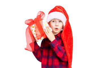Emotional surprised boy with wide open eyes and mouth holds and shakes a gift box with red bow. Child in Santa hat and pajamas with present looking at camera isolated on white background.
