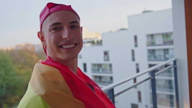 Young gay man with rainbow flag on the balcony turning around with a big smile. High quality 4k footage