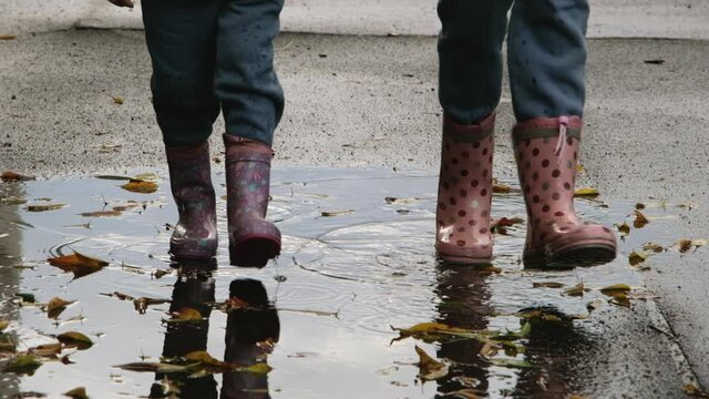 Two child girls in rubber boots are walking on wet asphalt covered with puddles and fallen leaves. Children and autumn. Full HD slow motion video.