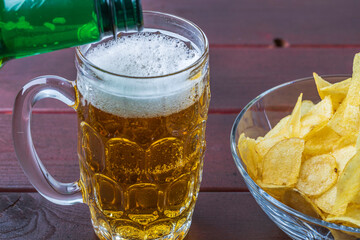 View of pouring beer into a mug and plates with potato chips isolated on wooden background. 