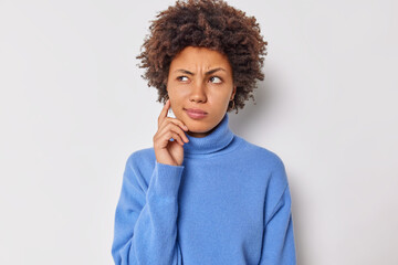 Fototapeta na wymiar Serious curly haired young woman considers something ponders about future plans keeps finger on cheek wears casual blue jumper isolated over white studio background. Let me think what to decide