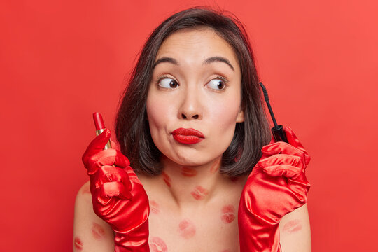 Portrait of beautiful brunette young woman holds lipstick and mascara looks away stands with naked body with traces of kiss applies cosmetics makes makeup poses against bright red background