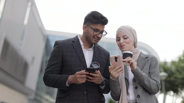 Arabian business couple having fun and talking together walking on the street during coffee break. Woman in hijab showing her smartphone screen to her colleague, sharing idea or news