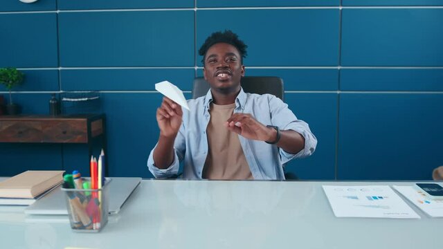 Optimistic young African American business man having fun in office folded paper into plane and launching, symbol of creativity at work, daydreaming about future vacation, career aspirations concept.