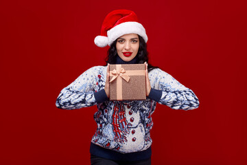 A cute young girl in a Christmas cap on a red background holds a gift box in front of her. Space for text.