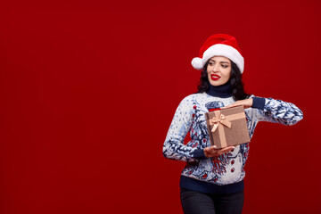 A pretty young woman in a cap demonstrates a New Year's gift on a red background. Emotional girl with red lipstick. Advertising banner for the new year.