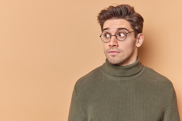 Photo of surprised adult handsome man looks with suspicion away notices something strange or shocking dressed casually poses against beige background blank copy space for your advertising content.