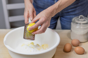 Woman is baking in domestic kitchen, indoors. Female hands grate lemon zest into dough. Horizontally.