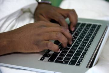Close-up of a black African man typing with his hands on a laptop computer sitting on a white bed