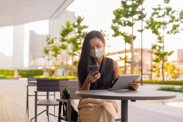 Woman sitting outdoors at coffee shop restaurant social distancing and wearing face mask to protect from covid 19 while using phone and digital tablet