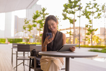 Portrait of beautiful Asian woman sitting outdoors at coffee shop restaurant smiling and using phone and digital tablet