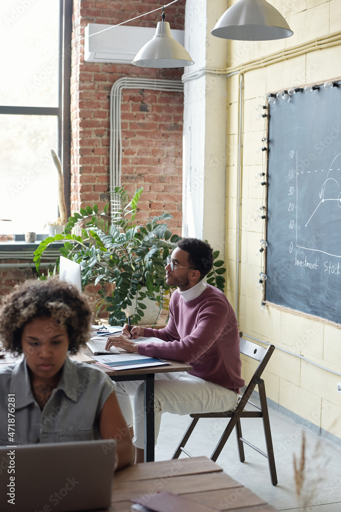 Poster young businessman sitting by workplace in front of computer monitor while his female colleague typin