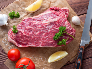 A piece of fresh raw beef lies on parchment surrounded by spices, herbs and vegetables. The knife is lying nearby. Top view, flat lay. Food composition